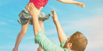 A father throws his daughter high into the air, in front of a light blue sky.